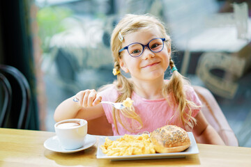 little smiling girl have a breakfast in a cafe. Preschool child with glasses drinking milk and eating scrambled eggs. Happy children, healthy food and meal.