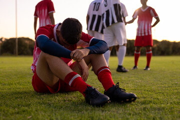 Wall Mural - Caucasian sad male athlete sitting on grassy land with team players in background at playground