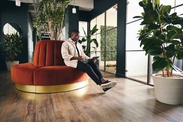 full length shot of casually dressed young man working on laptop in modern open plan office sitting 