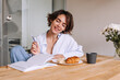 © Look! - Attractive brunette woman at the kitchen with notebook . In hands pencil looking and smiling at workbook with cup of coffee and croissant. Concept of lifestyle, working in home