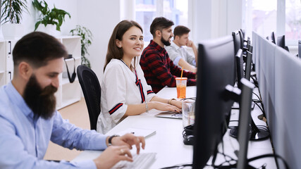 Junior female professional working in modern IT office. Group of young and experienced programmers and software developers sitting at desks working on computers.