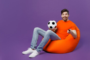 Wall Mural - Full body young fan man he wear orange t-shirt cheer up support football sport team hold soccer ball remote controller watch tv live stream sit in bag chair isolated on plain dark purple background.