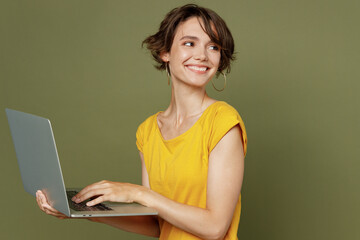 Side view young fun freelancer happy woman she 20s wear yellow t-shirt hold use work on laptop pc computer look aside on workspace area mock up isolated on plain olive green khaki background studio.