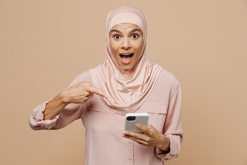 Young arabian asian muslim woman she wear abaya hijab pink clothes use point finger on mobile cell phone isolated on plain pastel beige background. People uae middle eastern islam religious concept.