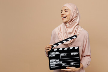 Young arabian asian muslim woman she wear abaya hijab pink clothes hold classic black film making clapperboard look aside isolated on plain pastel beige background. People uae islam religious concept.