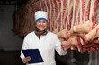 Meat-packing plant worker in front of butchered carcasses. Meat production. A professional butcher between rows of pork carcasses, looking at the camera, posing. 