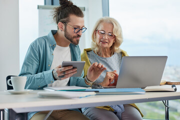 A young professor sits next to a female senior student and helps her to master web design.