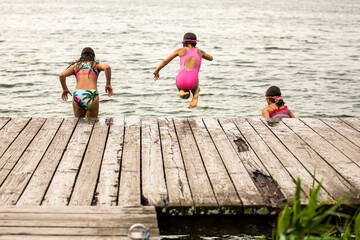 Wall Mural - Back view of three girls jumping into water of lake from wooden pier on cloudy summer day