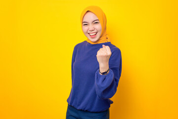 Wall Mural - Excited young Asian Muslim woman dressed in casual sweater celebrating success with raised fist isolated over yellow background
