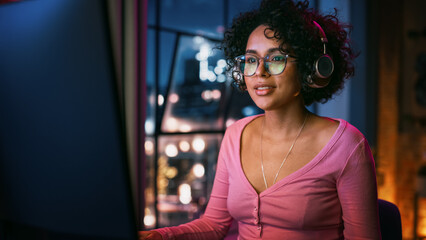 Fototapete - Beautiful Multiethnic Black Woman in Headphones Using Computer in Stylish Loft Apartment in the Evening. Creative Female Smiling, Browsing Videos on Social Media. Urban City View from Big Window.