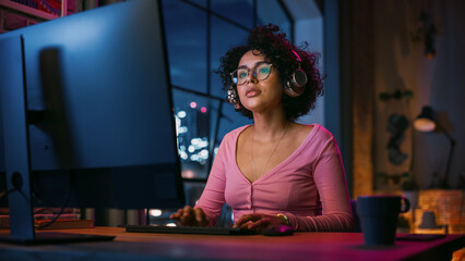 Fototapete - Attractive Multiethnic Latin Woman in Headphones Using Computer in Stylish Loft Apartment in the Evening. Creative Female Smiling, Browsing Videos on Social Media. Urban City View from Big Window.