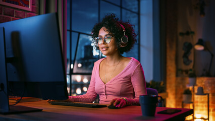 Wall Mural - Young Beautiful Black Woman in Headphones Using Computer in Stylish Loft Apartment in the Evening. Creative Female Smiling, Browsing Videos on Social Media. Urban City View from Big Window.