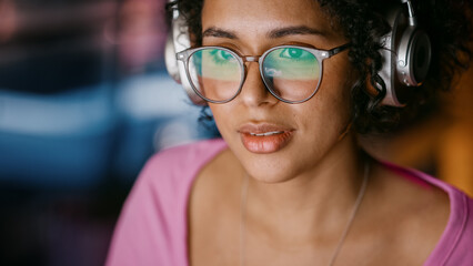 Wall Mural - Close Up Portrait of Beautiful Multiethnic Black Woman in Headphones Using Computer in Stylish Loft Apartment in the Evening. Creative Female Smiling, Browsing Videos on Social Media.