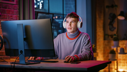 Wall Mural - Young Handsome Man in Headphones Using Computer in Stylish Loft Apartment in the Evening. Creative Male Smiling, Working from Home, Browsing Videos on Social Media. Urban City View from Big Window.