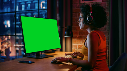 Fototapete - Young Beautiful Female in Headphones Working from Home on Desktop Computer with Green Screen Mock Up Display. Creative woman Checking Social Media, Browsing Internet. Urban City View from Big Window.