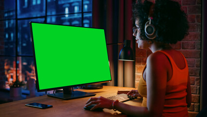 Fototapete - Young Beautiful Female in Headphones Working from Home on Desktop Computer with Green Screen Mock Up Display. Creative woman Checking Social Media, Browsing Internet. Urban City View from Big Window.