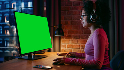 Fototapete - Young Beautiful Female in Headphones Working from Home on Desktop Computer with Green Screen Mock Up Display. Creative woman Checking Social Media, Browsing Internet. Urban City View from Big Window.