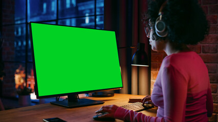 Fototapete - Young Beautiful Female in Headphones Working from Home on Desktop Computer with Green Screen Mock Up Display. Creative woman Checking Social Media, Browsing Internet. Urban City View from Big Window.