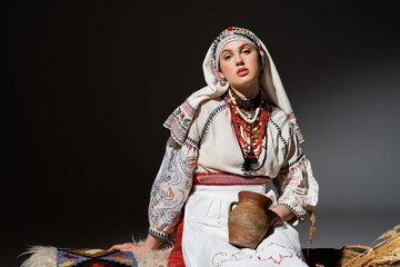 Wall Mural - young ukrainian woman in traditional clothing with ornament holding clay pot and sitting on bench on black.