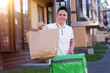 © Евгений Шемякин - portrait of young handsome happy courier guy, food delivery man with green thermo box for food delivering food outdoors in the yard on buildings background at summer day in cap and uniform.