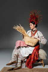 Wall Mural - full length of pretty ukrainian woman in traditional clothes and red wreath with flowers holding bag with wheat on dark grey.