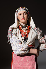 Wall Mural - young ukrainian woman in traditional shirt with ornament and red beads posing with hand on hip on black.