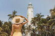 Back view of romantic woman traveler in dress and straw hat visiting famous landmarks of Sri Lanka country standing and looking on Dondra lighthouse. High quality photo.