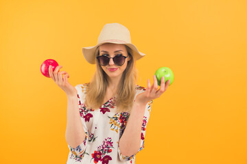 Temptation to eat fruit. Studio shot of a European girl in her mid 20s wearing floral blouse, sunglasses, and a hat holding up two apples - one apple is red, the other one is green. Orange background