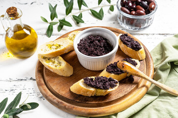 Olive pate in bowl and spreader on bread slices on wooden cutting board. 