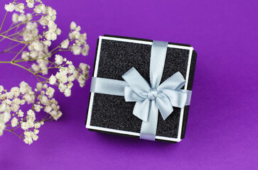 Black gift box with a white ribbon on a purple background with a bouquet of gypsophila