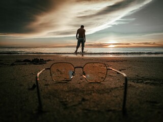 Closeup of glasses on the beach with a standing man in the background of the sunset.