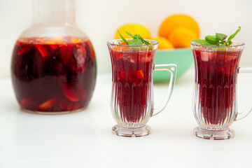 Full jar and two glasses of homemade red wine sangria cocktails ice and mint inside, with a bowl of orange, lemon, apricot background on a white table. copyspace