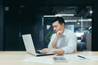 © Liubomir - A young handsome man, an Asian businessman, is working with a laptop, typing. Sitting at a desk in a modern office