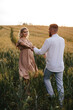 © VikaNorm - A pregnant woman in a beautiful long dress walks through the field with her husband in a wheat field at sunset. Future parents.