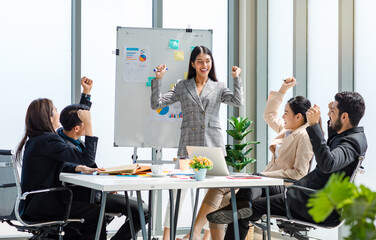 Asian businesswoman presenter in formal suit standing smiling holding pen pointing at graph chart document on whiteboard presenting information to multinational male female colleagues in meeting room