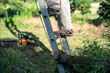 Closeup of man's feet in work boots climbing a ladder with chainsaw background, his shadow, and cut tree branches in a sunny yard with green grass