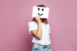 Cute woman covering her face using white paper with smile sign on pink background