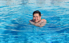 Asian Man In The Swimming Pool. A Young Man Swimming In The Pool And Holding A Ball. Active Resting. Asian Male Squinting Eyes From The Sun.