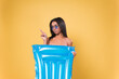 Young African American woman wearing bikini, holding an air mattress points to the side on yellow background.