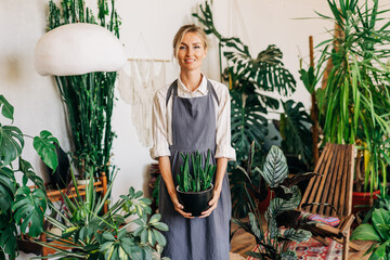 Wall Mural - A young woman owner of a greenhouse stands in an apron holding a potted plant.