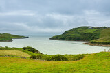 Fototapeta  - A wide bay with hills covered with green trees under a thick cloudy sky. The sandy beach of a cove-shaped bay with lush green grass near the edge of the ocean.
