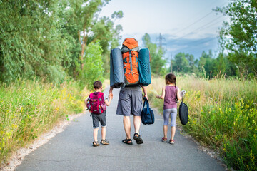 Wall Mural - Back view of father carrying backpack with son and daughter walking along way in forest on summer sunset outdoors