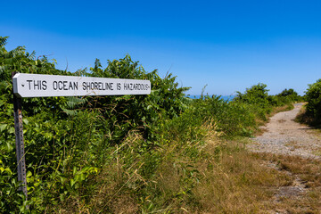 A narrow walkway along the beach with a sign saying This Ocean Shoreline Is Hazardous 