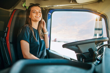 Portrait of beautiful young woman professional truck driver sitting in a big truck, looking at camera and smiling. Inside of vehicle. People and transportation concept.