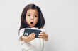 © Tatiana - a cute, funny, little girl in a white t-shirt stands on a light background with a fashionable smartphone in her hand and takes a selfie with her mouth wide open.