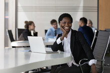 Happy Young African American Business Woman Corporate Portrait. Joyful Female Business Leader, Employee Posing At Laptop, Sitting At Work Table In Office, Looking At Camera, Smiling, Touching Chin