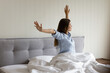 Young rested after healthy night sleep woman wake up do stretching exercises sit in bed, feeling refreshed satisfied raised hands enjoy early morning at home or hotel. Good morning, comfort concept