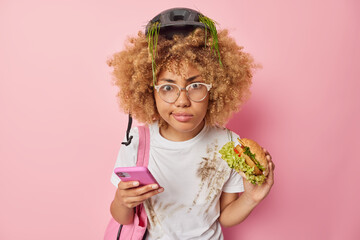 Serious curly haired woman focused into camera eats burger uses smartphone prefers fast food wears protective helmet and casual white t shirt isolated over pink background. Unhealthy eating.