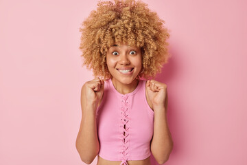 Wall Mural - Excited curly haired celebrates something triumphs over something bites lips awaits for something wears top poses against pink background winning and rejoicing await for announcement of results