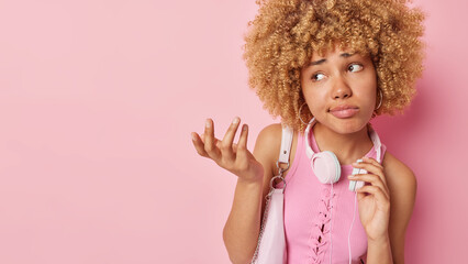 Wall Mural - Hesitant clueless young woman with curly hair shrugs shoulders feels confused carries bag wears headphones around neck dressed in t shirt isolated over pink background copy space for your promo